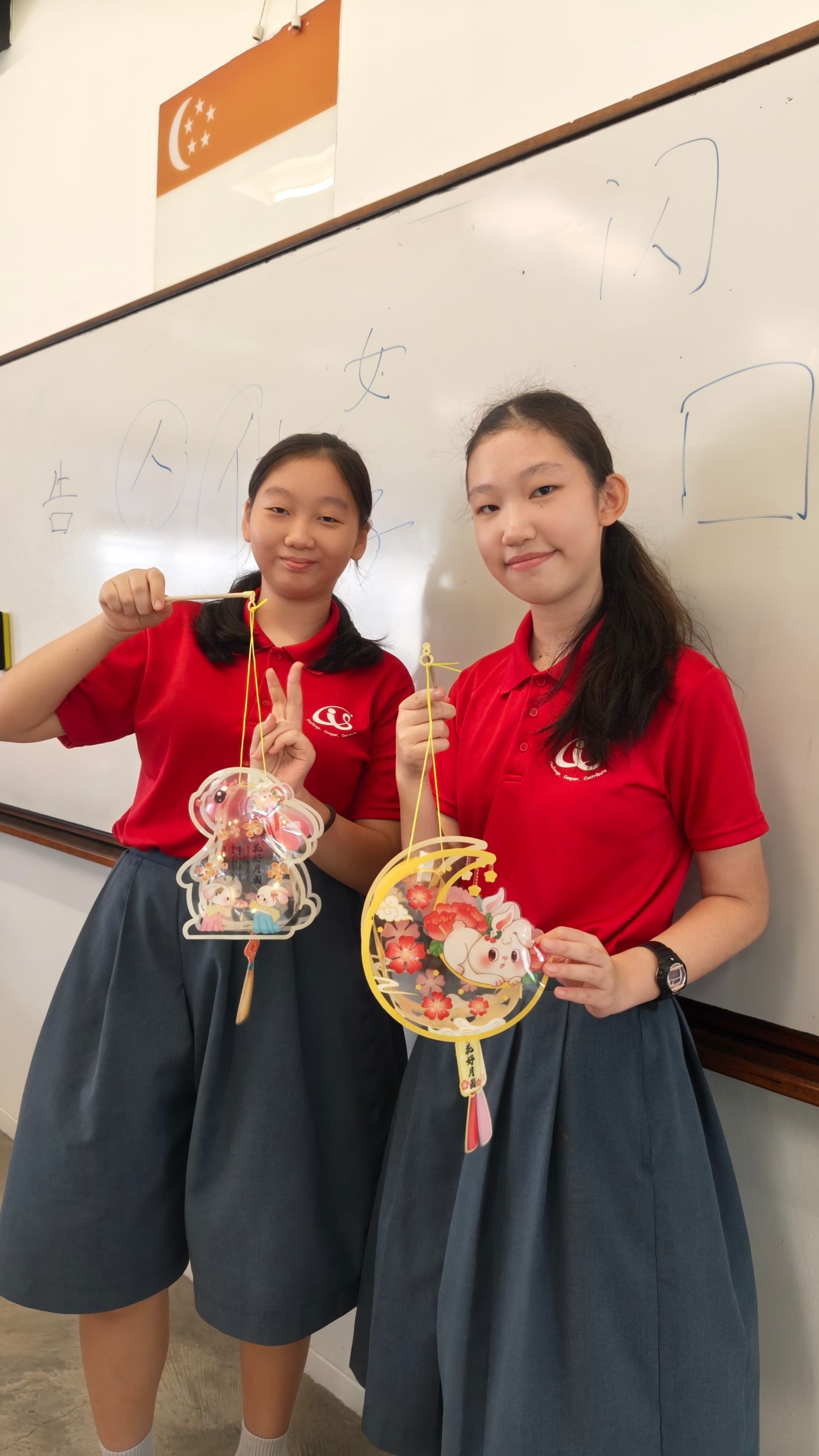 Two female students in red school uniforms stand in front of a whiteboard with Chinese characters written on it. They smile and hold up handmade Mid-Autumn Festival lanterns — one rabbit-shaped and one circular moon-shaped lantern with floral decorations. One student flashes a peace sign.